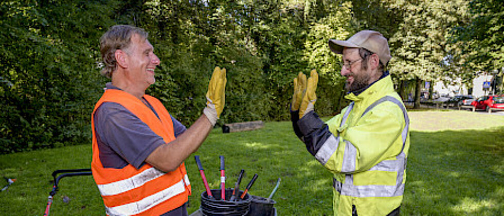 Zwei Bauarbeiter mit Warnkleidung geben sich draußen auf einer Wiese lachend und freudig einen High-Five. Im Hintergrund sind Werkzeuge und ein Schubkarren zu sehen.