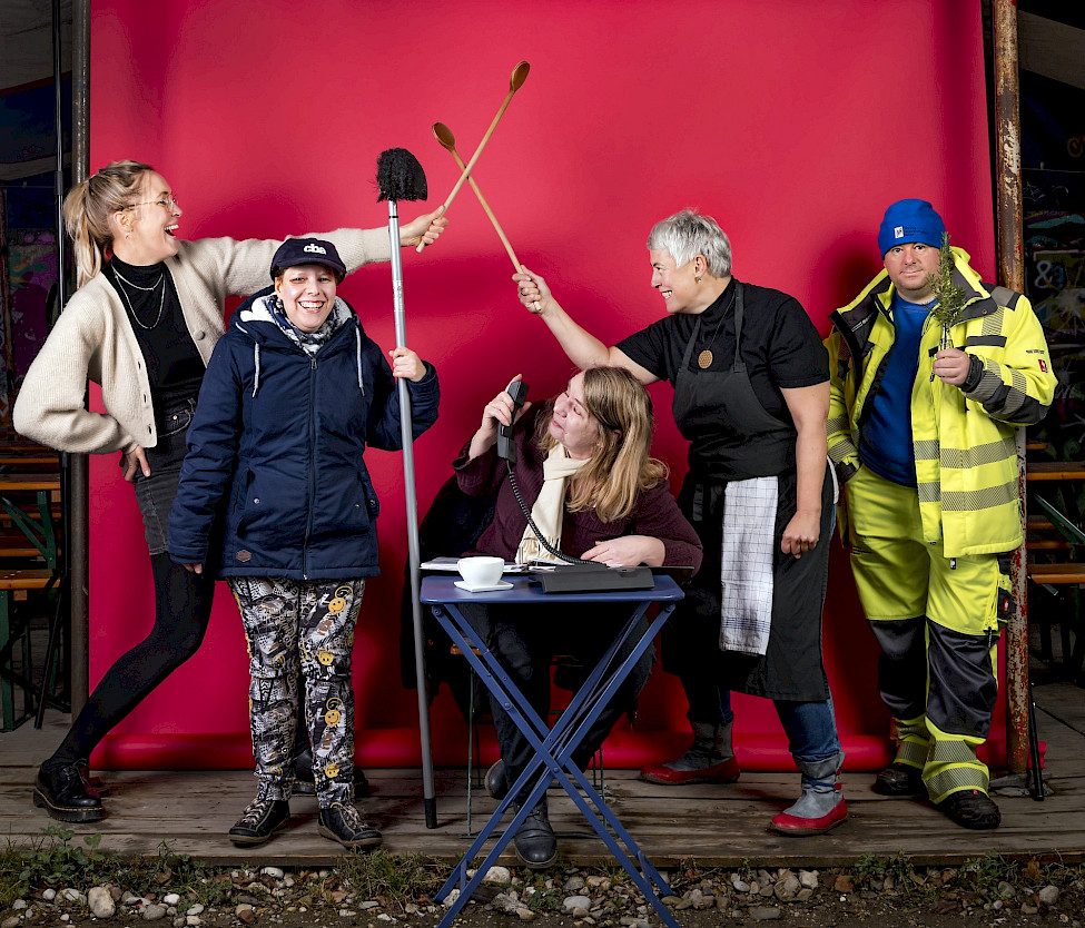 Fünf Personen mit unterschiedlichen Arbeitskleidungen posieren vor rotem Hintergrund. Zwei halten Kochlöffel, eine einen Toilettenbürstenkopf, eine sitzt am Tisch mit Mikrofon, und eine trägt Warnschutzkleidung. Alle wirken fröhlich und sind aktiv miteinander verbunden.